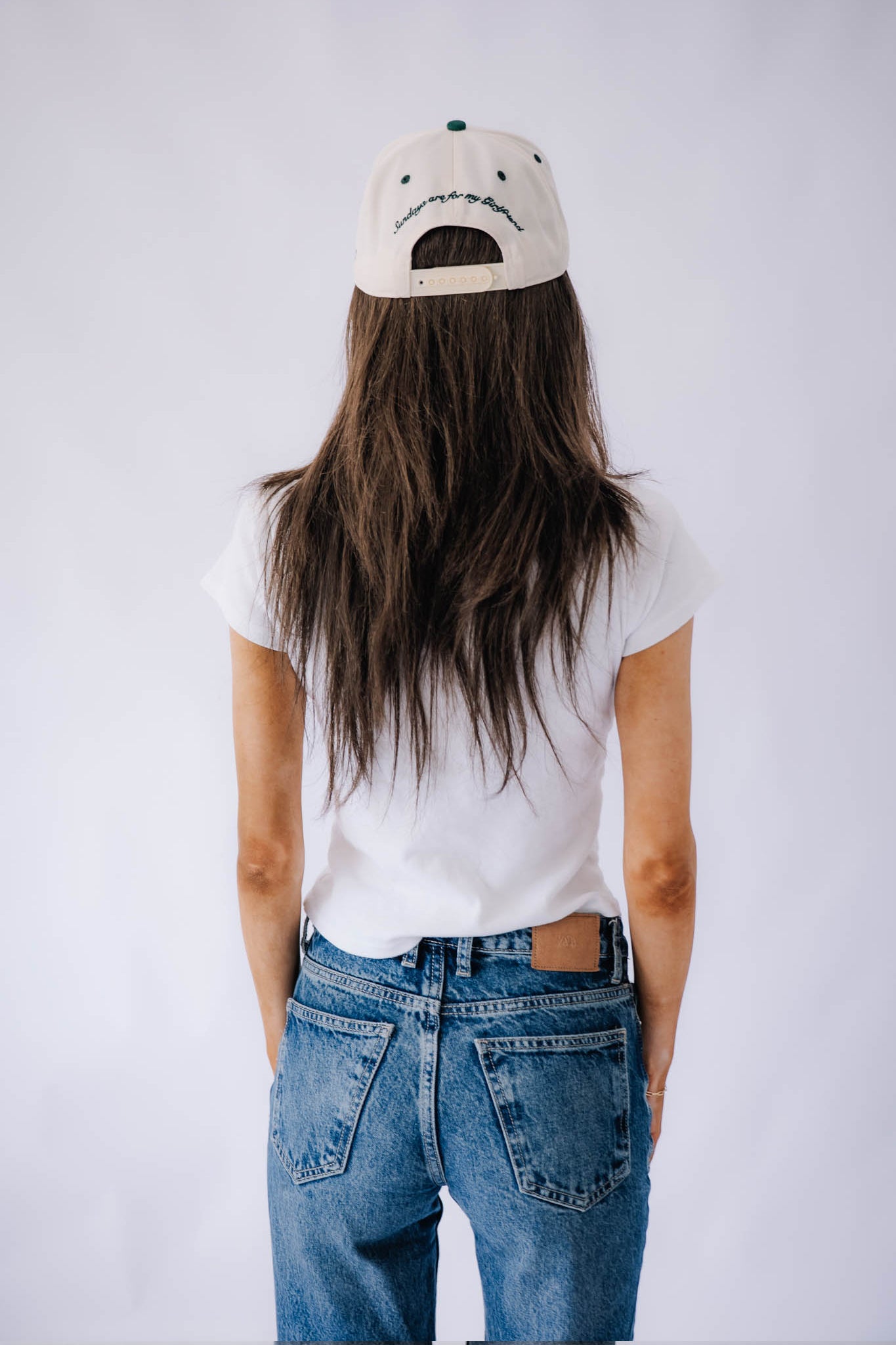 Person wearing a white t-shirt and blue jeans with a green forest cap on a plain background