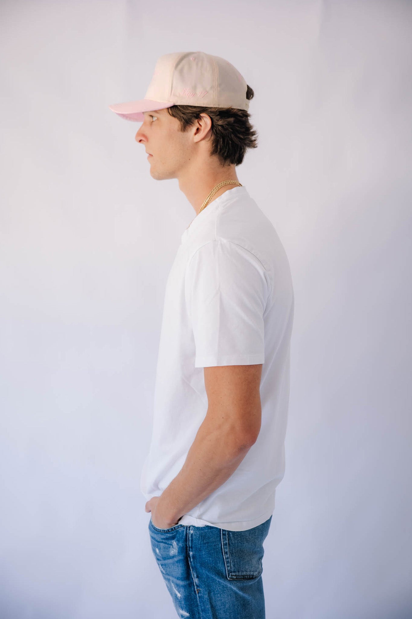 Man wearing a white t-shirt, blue jeans, and a pink cap against a plain background