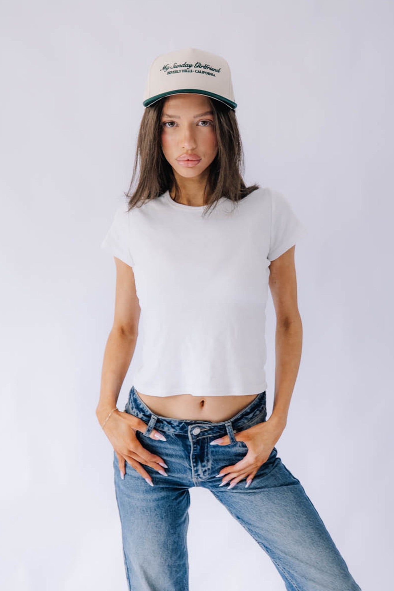 Woman wearing a white t-shirt and blue jeans with a green forest cap on a plain background