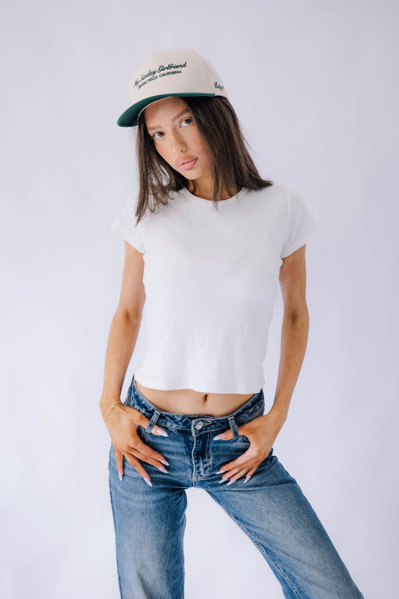 Person wearing a white t-shirt, blue jeans, and a green forest cap on a plain background