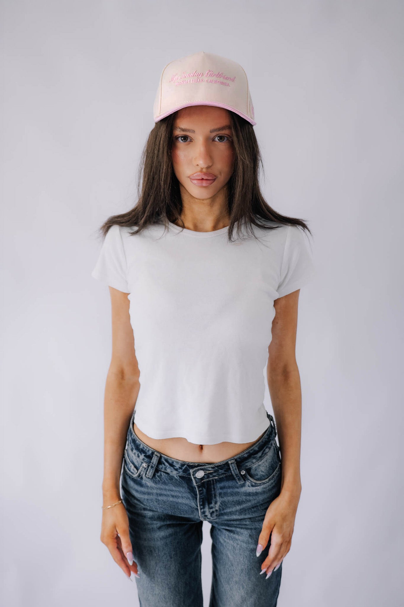 Person wearing a white t-shirt, blue jeans, and a pink cap on a plain background