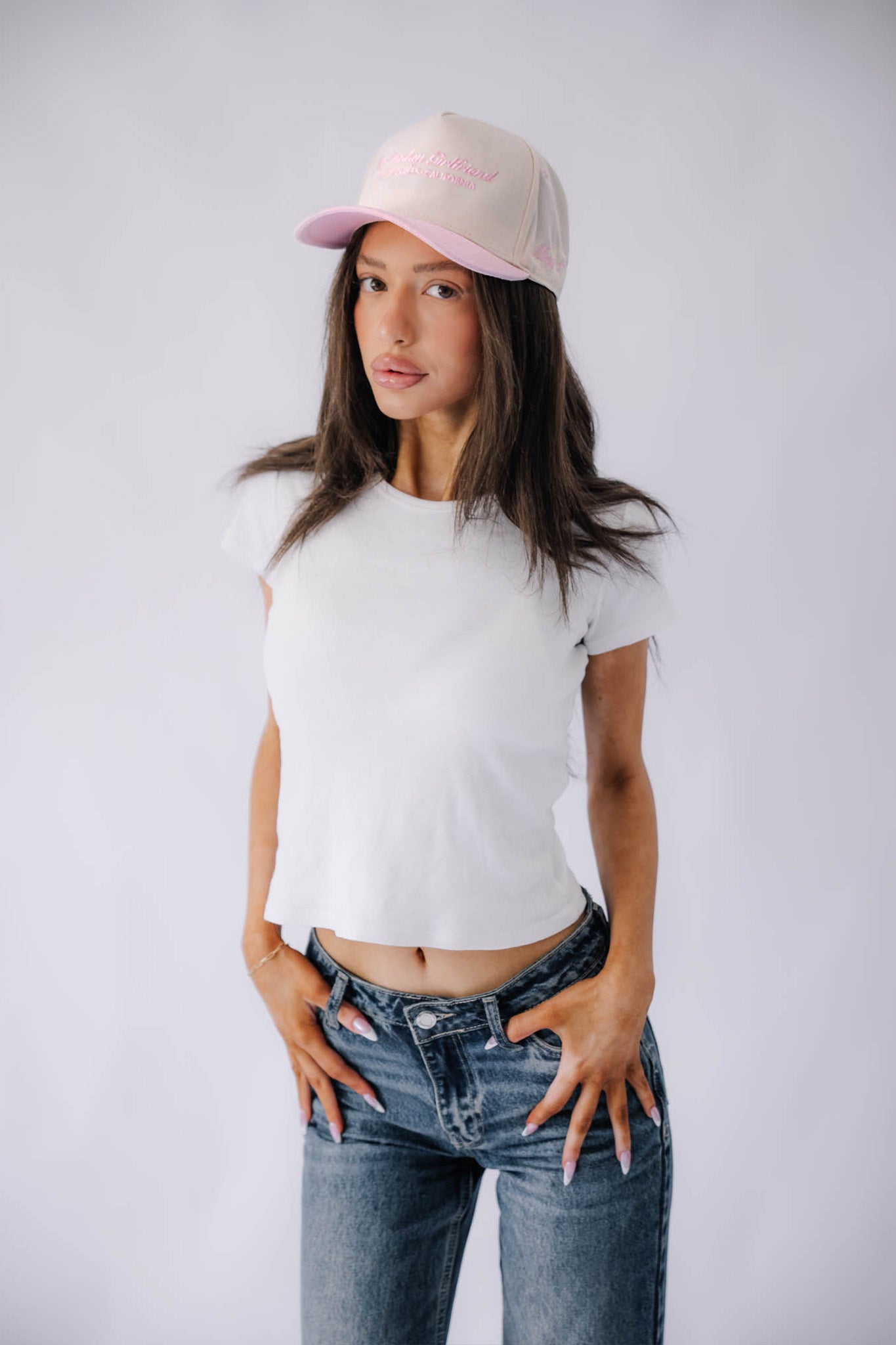 Woman wearing a white t-shirt, blue jeans, and a pink cap on a plain background